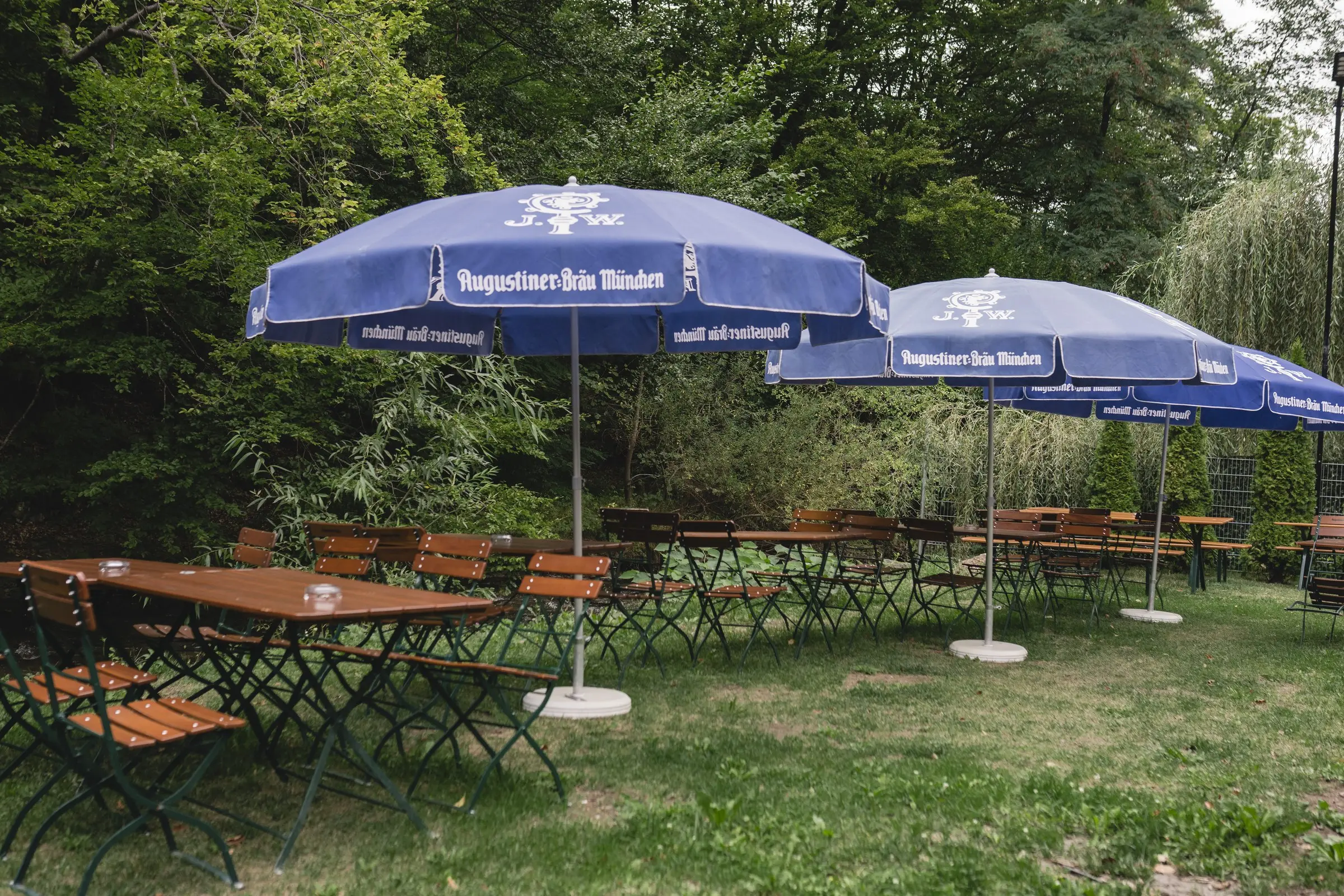 Mese de lemn sub umbrele Augustiner, pe terasa înconjurată de verdeață
